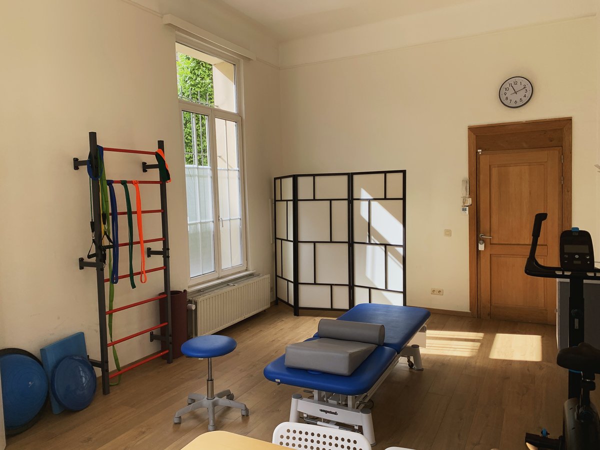 Salle de soins du cabinet de kinésithérapie Broxolle Care à Ixelles, Bruxelles — table de traitement, espalier et paravent devant une grande fenêtre lumineuse